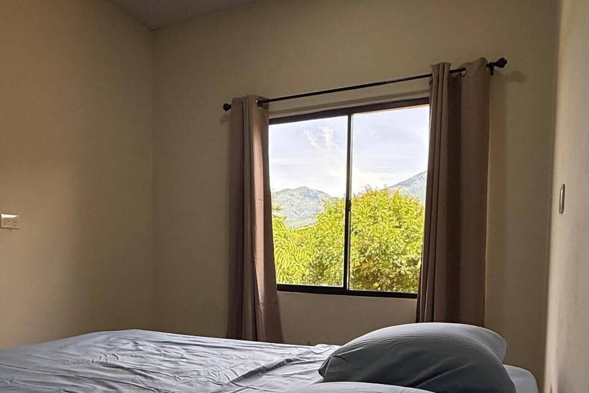 A bedroom with a bed, a window with curtains, and a view of greenery and mountains.