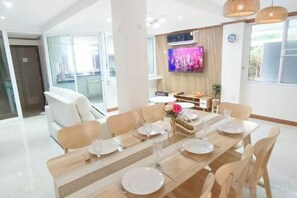 Un comedor con una mesa larga dispuesta para seis personas, rodeada de sillas, con un televisor montado en la pared y un ventanal que proporciona luz natural.