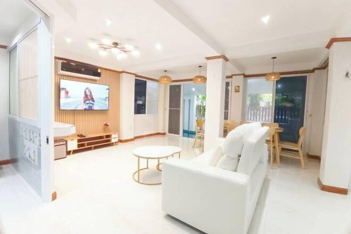 A modern living room with a white sofa, a TV mounted on the wall, and a dining area with a table and chairs.
