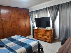 A bedroom with a bed, wooden wardrobe, TV on a dresser, and grey curtains.