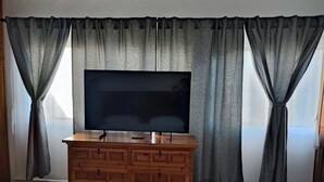 Ett rum med en byrå i trä, en platt-TV ovanpå och grå gardiner.