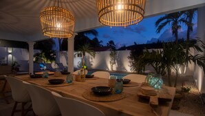 An outdoor dining area with a wooden table, white chairs, and hanging wicker lamps. - *NEW* Cozy Casa! Tasteful family villa with private pool! (Oranjestad)