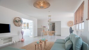 A modern living room with a sofa, coffee table, and TV. There is a dining area with chairs and a table, and a large window with blinds. - *NEW* Cozy Casa! Tasteful family villa with private pool! (Oranjestad)