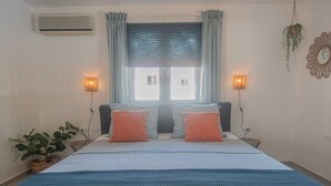 A bedroom with a bed, two bedside lamps, a window with curtains, a wall-mounted air conditioner, and a potted plant. - *NEW* Cozy Casa! Tasteful family villa with private pool! (Oranjestad)