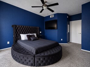 A bedroom with a tufted headboard, a round ottoman, a ceiling fan, a flat-screen TV, and a white door.