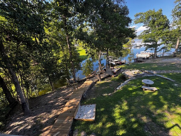 Eine Holzterrasse führt zu einem Steg an einem See, umgeben von Bäumen und Grünflächen.