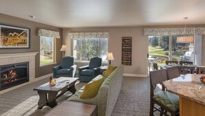 A living room with a fireplace, a sofa, two armchairs, a dining table with chairs, and a view of a garden through large windows. - Enjoy Oktoberfest in this Charming 2-bedroom condo in fabulous Leavenworth (Leavenworth)