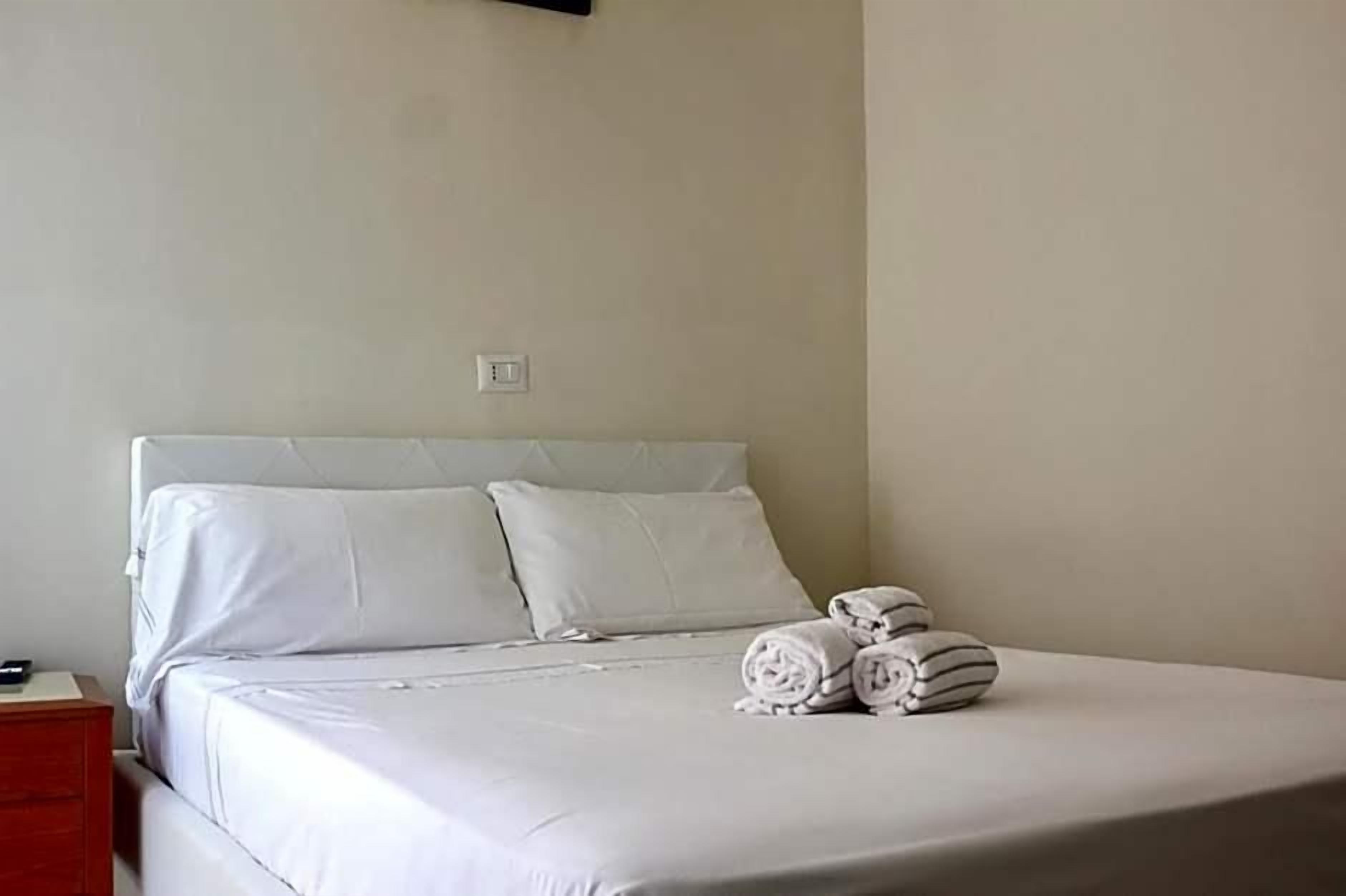 A neatly made bed with white linens and pillows, a bedside table with a lamp, and rolled towels on the bed.