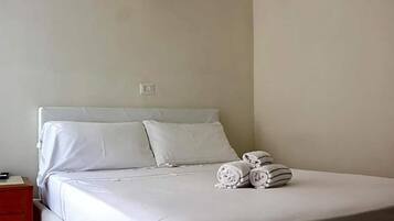 A neatly made bed with white linens and pillows, a bedside table with a lamp, and rolled towels on the bed.