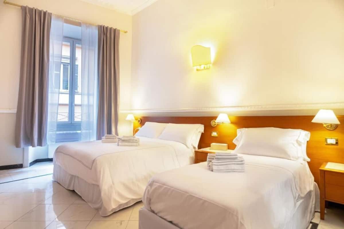 A hotel room with two beds, a window with curtains, and wall-mounted lamps.