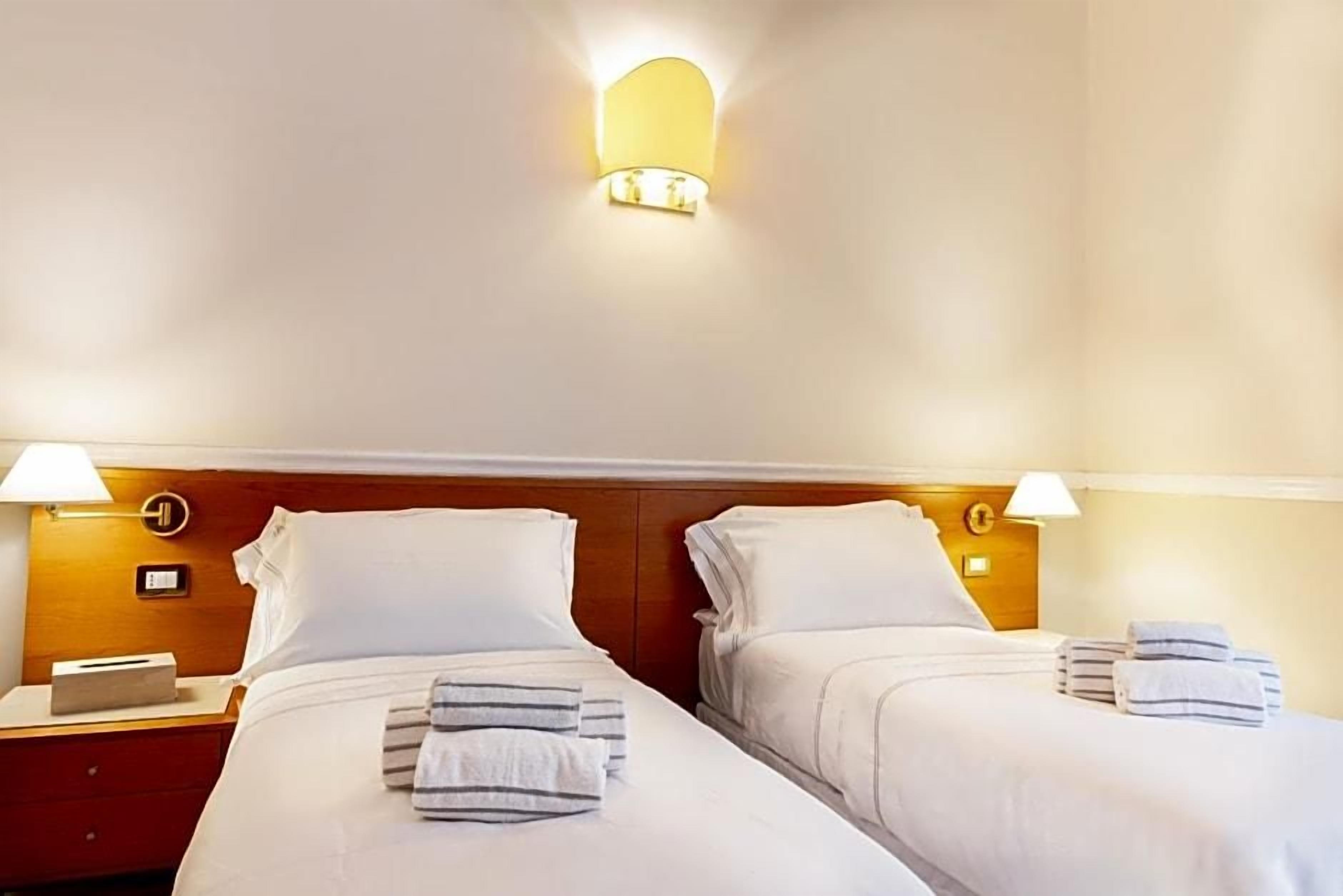 A hotel room with two beds, each with a nightstand, a lamp, and towels.