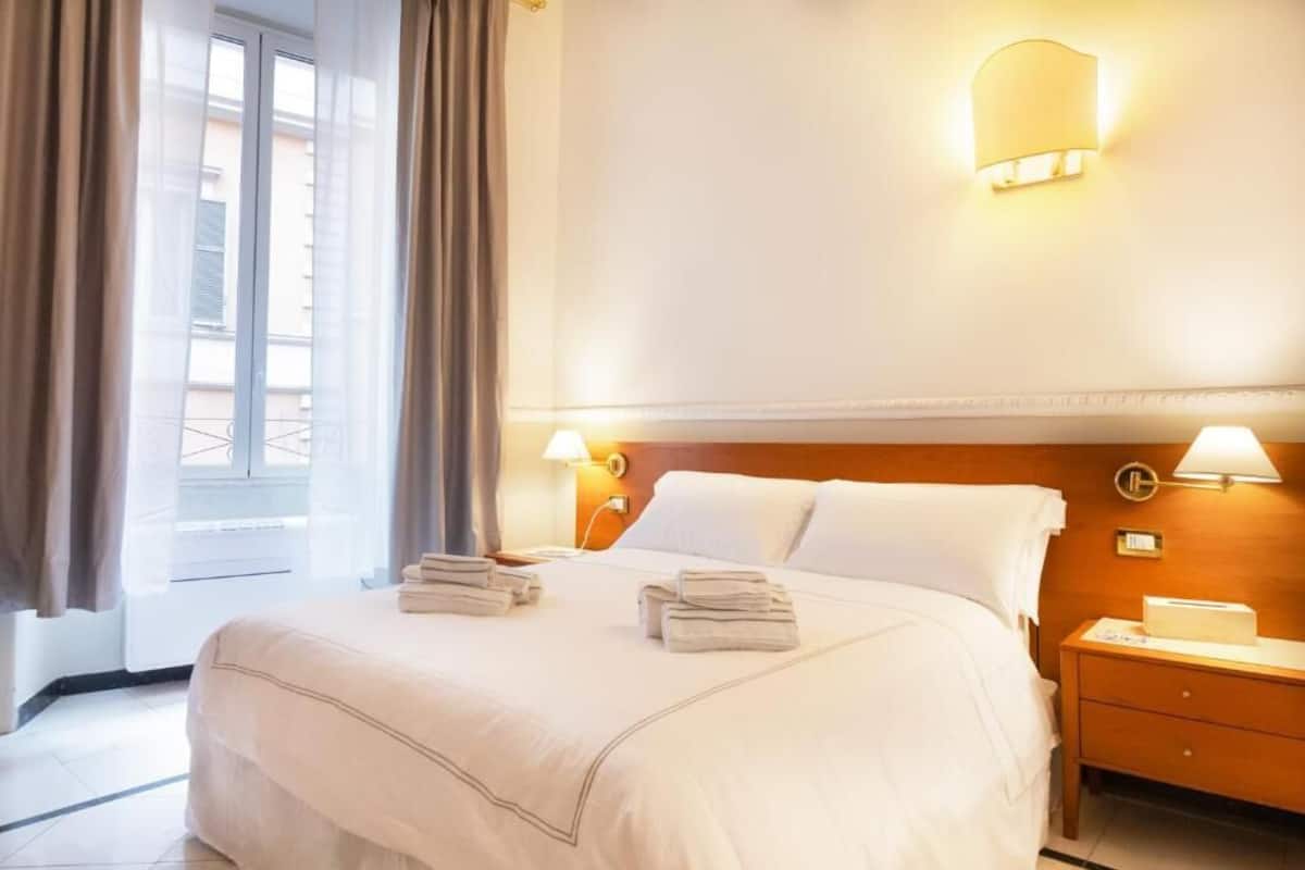 A hotel room with a bed, two bedside tables, a nightstand, a window with curtains, and wall-mounted lamps.