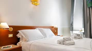 A hotel room with a neatly made bed, two bedside lamps, a small bedside table with a book, and a window with curtains.
