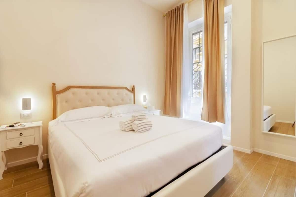 A neatly made bed with a wooden headboard, a bedside table with a lamp, and a window with curtains.
