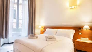 A hotel room with a bed, two bedside tables, a nightstand, a window with curtains, and wall-mounted lamps.
