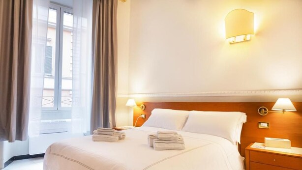 A hotel room with a bed, two bedside tables, a nightstand, a window with curtains, and wall-mounted lamps. - Hotel Domus Rome (Rome)