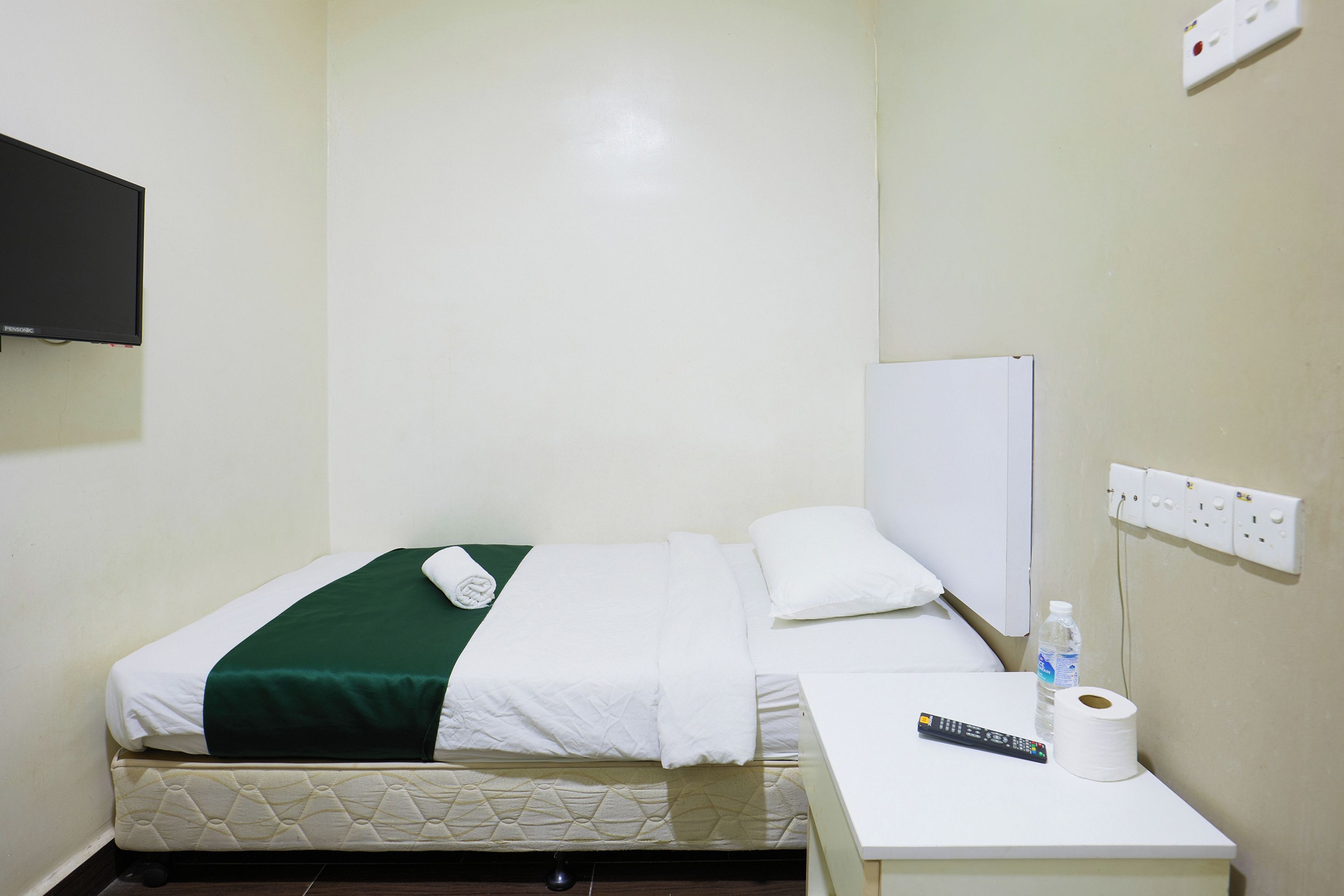 A hotel room with a bed, a bedside table with a remote control, a roll of toilet paper, and a water bottle.