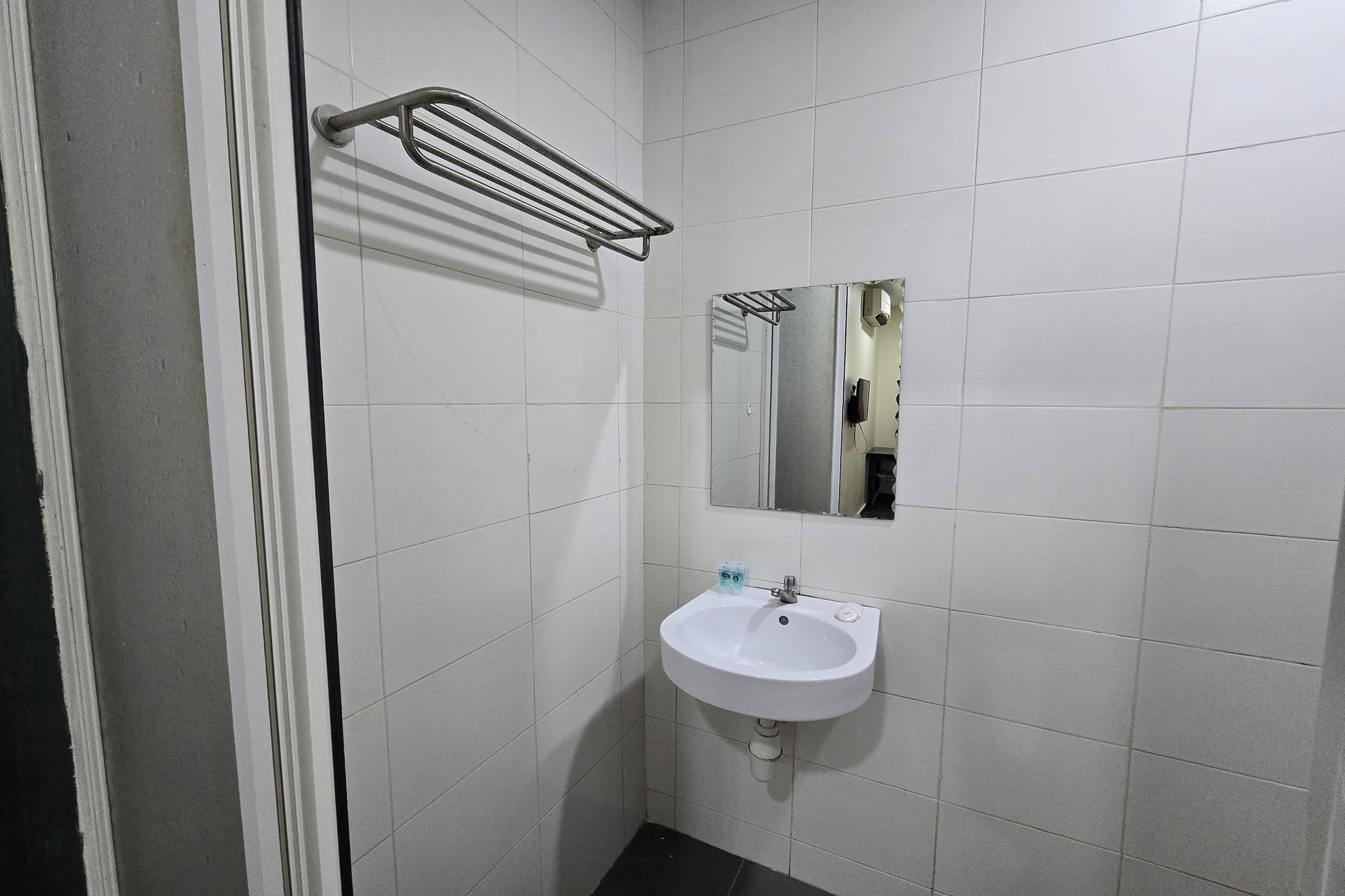 Une salle de bain avec des murs recouverts de carreaux blancs, un lavabo blanc avec un miroir au-dessus, et un porte-serviettes.