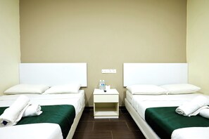 Habitación de hotel con dos camas, cada una con ropa blanca y colchas verdes. Se observa una mesita de noche pequeña con un control remoto, una botella de agua y una caja de pañuelos.