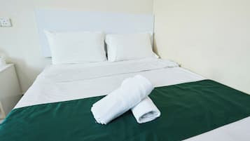 Una cama bien hecha con sábanas blancas, una colcha verde y dos toallas blancas enrolladas.