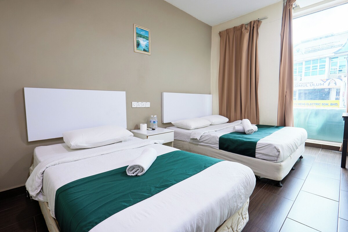 Une chambre d’hôtel avec deux lits, une fenêtre avec des rideaux, une table de chevet avec une bouteille et une boîte de mouchoirs, et une prise électrique fixée au mur.