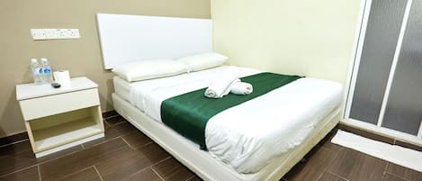 A neatly made bed with a green blanket and white linens, a bedside table with a remote control and bottled water, and a power outlet on the wall.