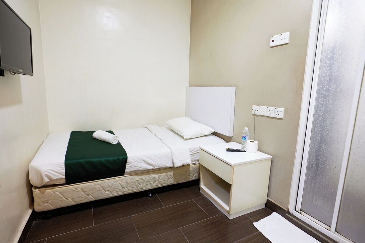 Ein kleines, ordentlich eingerichtetes Hotelzimmer mit einem Bett, einem Nachttisch mit Fernbedienung und einer Wasserflasche, einem an der Wand montierten Fernseher und einem Fenster mit Blick ins Freie.