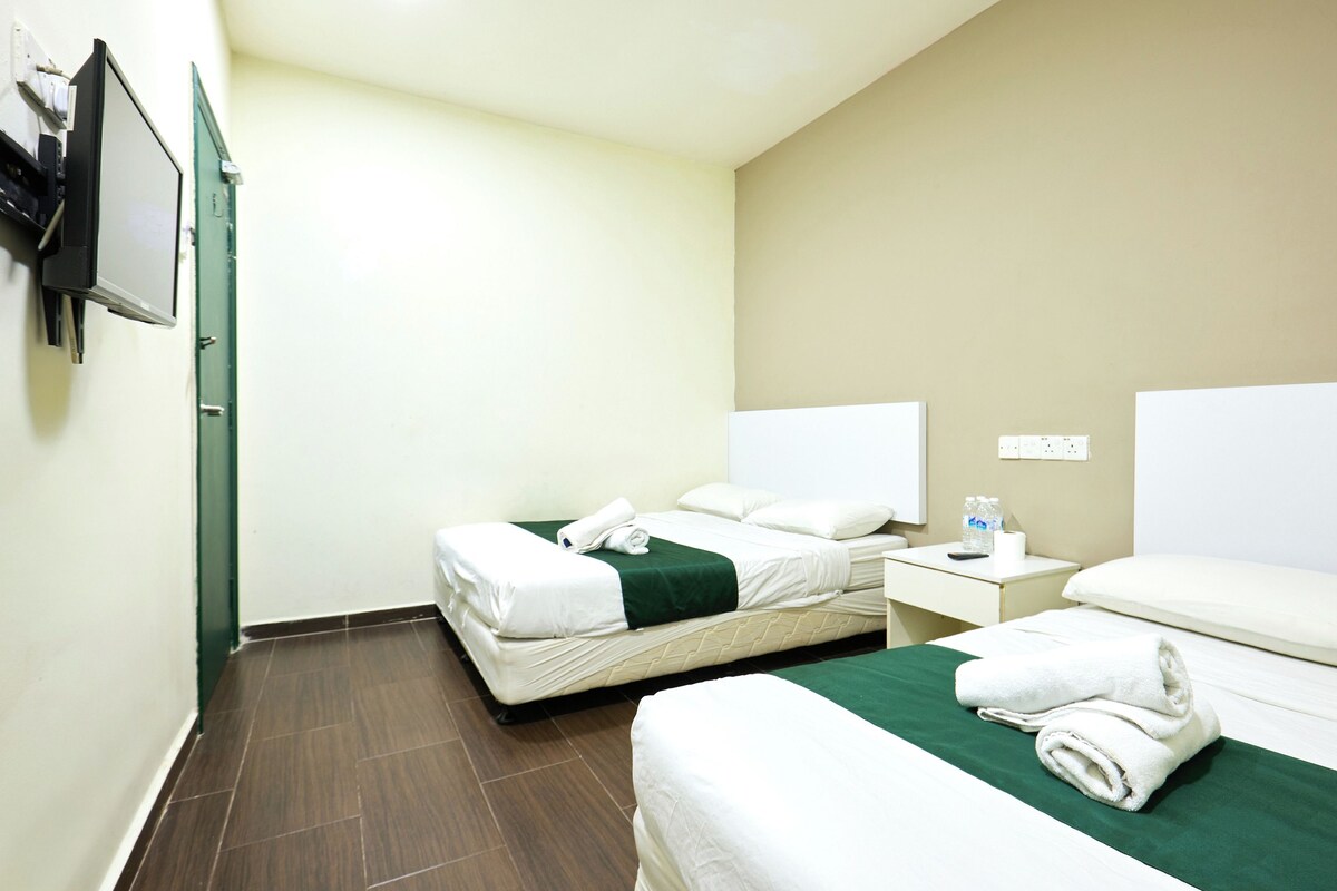 Habitación de hotel con dos camas, una colcha verde, ropa de cama blanca, una mesita de noche con una botella y un teléfono, y un televisor montado en la pared.