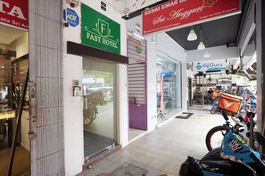 Une zone commerciale avec l’entrée d’un hôtel économique, une enseigne indiquant « Kedai Emas Sri Anggun » et une moto garée à l’extérieur.