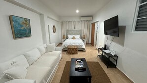 A modern, well-lit bedroom with a white bed, a white sofa, a coffee table, and a TV on a stand.