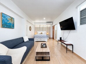A modern hotel room with a bed, a sofa, a coffee table, a TV, and a window with blinds.