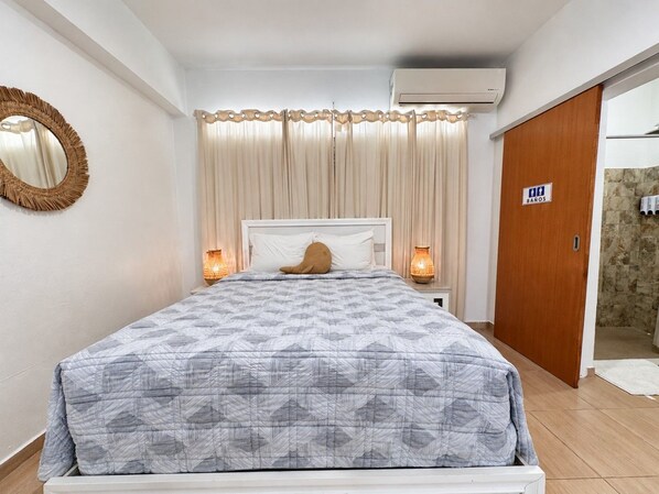 A hotel room with a bed, curtains, a wall-mounted mirror, a wooden door, and an air conditioning unit.