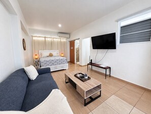 A modern hotel room with a bed, a sofa, a coffee table, a TV, and a window with blinds.
