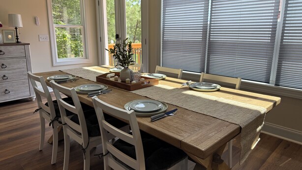 A dining room with a wooden table set for six, white chairs, and a view of the outdoors through large windows. - Charming 2-bedroom house-Walk to Helen-Your Perfect Getaway (Helen)
