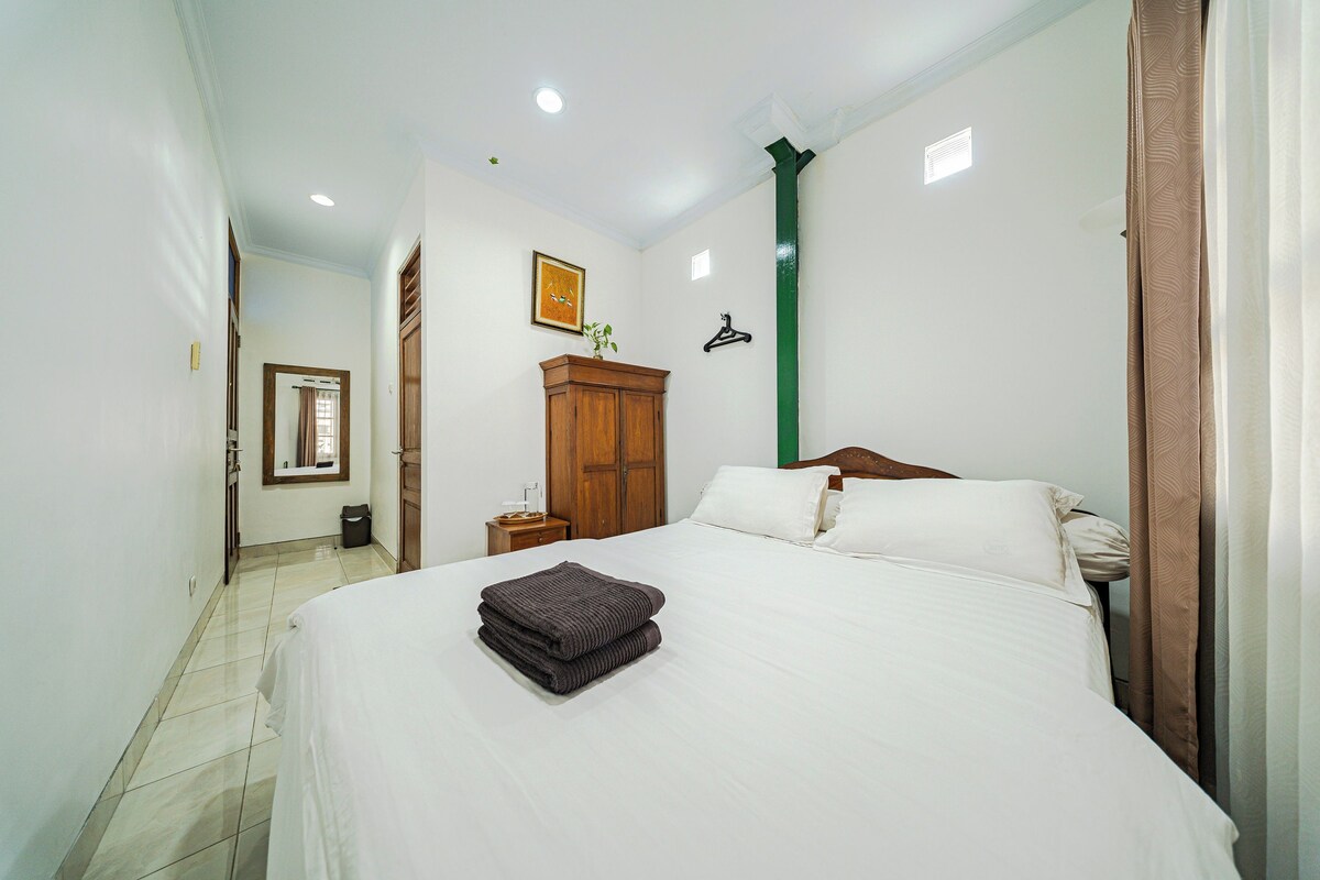 Un dormitorio con una cama, una mesita de noche, un armario de madera, un pilar verde y una ventana.