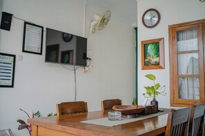 A dining area with a wooden table, chairs, a clock, a television, and a window with curtains. - Omah Gerjen 29 By Zuzu (Yogyakarta)