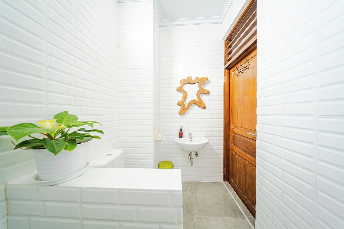 Un baño con azulejos blancos tipo metro, una puerta de madera, un lavamanos blanco y una planta en maceta.