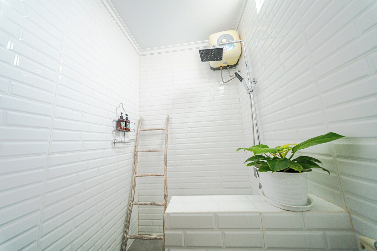 Baño con paredes de azulejos blancos, ducha, escalera, planta en maceta y estante con artículos de tocador.