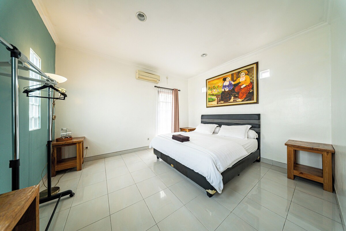 Una habitación de hotel con cama, mesitas de noche, un cuadro, una ventana con cortinas y un radiador.