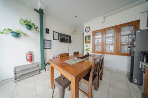 Una mesa de madera con sillas, un extintor, un taburete pequeño, un ventilador de pared y un refrigerador.