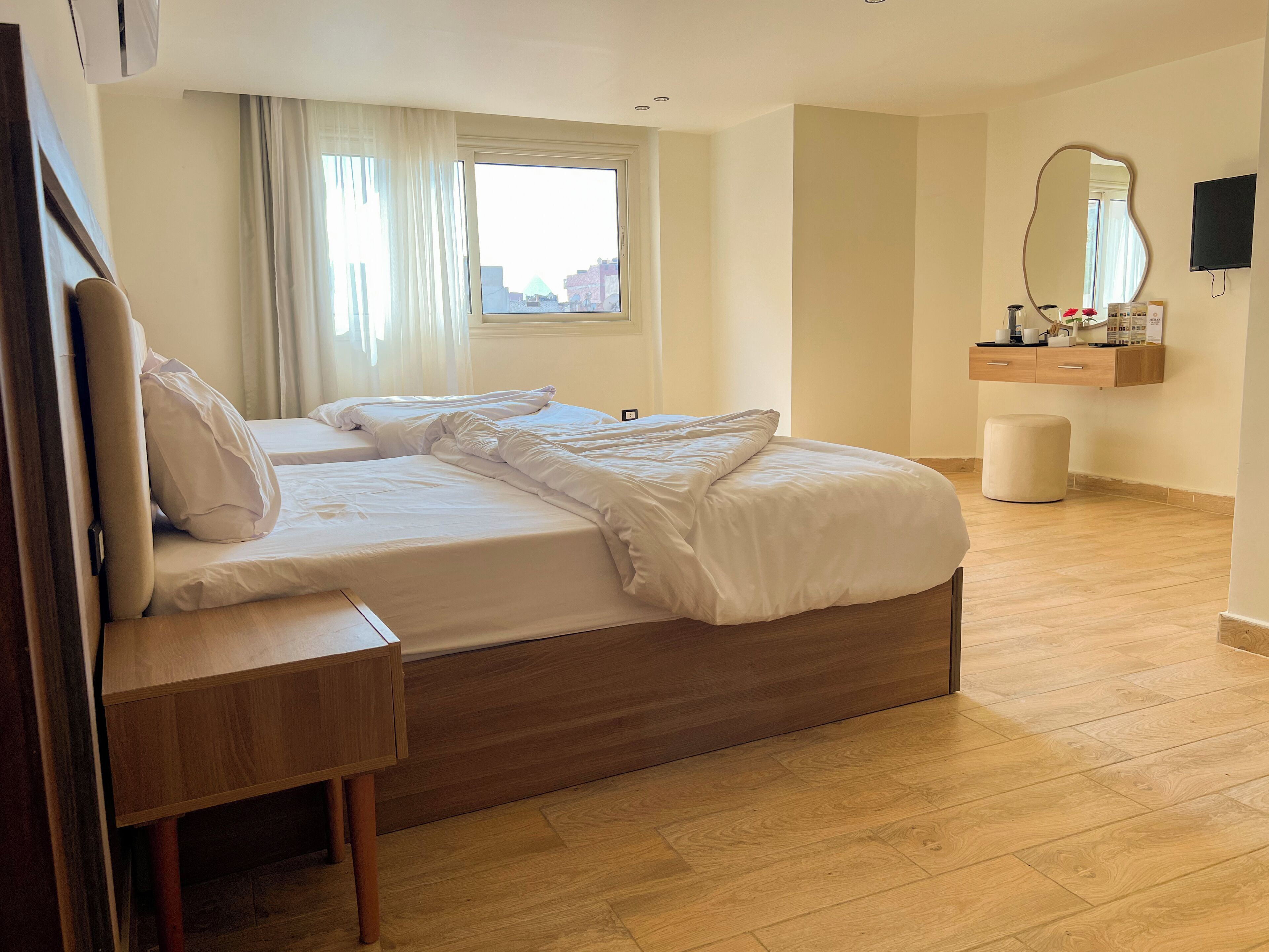A hotel room with a wooden bed, a wooden nightstand, a mirror, a TV, and a window with curtains.