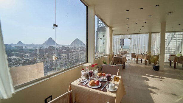 A dining area with a table set for breakfast, a view of pyramids, and a modern interior design. - Merak Pyramids View Hotel (Giza)