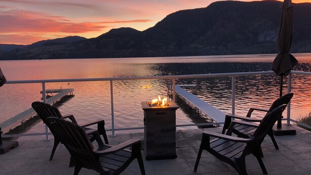 Eine Terrasse mit zwei Stühlen und einem Feuerkorb mit Blick auf einen ruhigen See, im Hintergrund Berge.