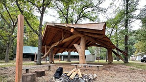 Een rustiek houten paviljoen met schuin dak, omgeven door bomen en een picknicktafel.