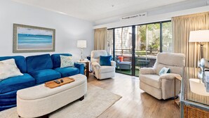 Un salon spacieux avec un canapé bleu, deux fauteuils et une table basse. De grandes fenêtres offrent une vue sur l’extérieur.