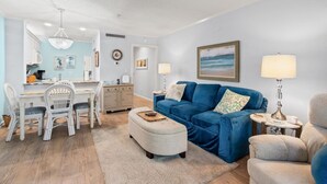Un salon avec un canapé bleu, un coin repas avec des chaises blanches et une cuisine.