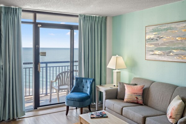 Quarto com vista para o oceano, uma poltrona azul, um sofá cinza, uma mesa pequena com abajur e um quadro na parede.