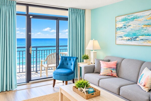 A room with a view of the ocean, a blue armchair, a grey sofa, and a painting on the wall.