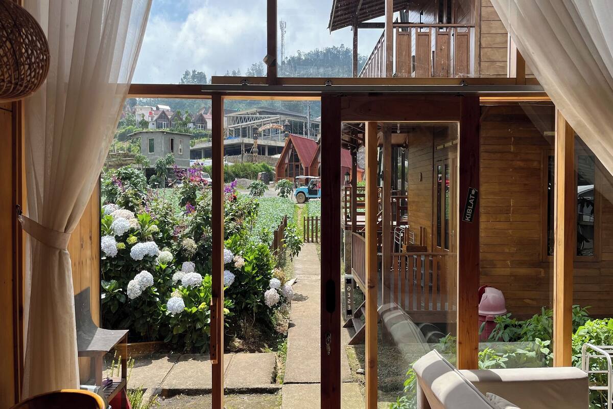 A view from a room with a large window, looking out to a garden with hydrangeas and a blue car parked outside.