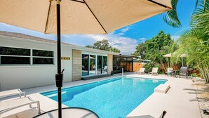 Una piscina con sillones de descanso y sombrillas, rodeada por una casa blanca y plantas tropicales.